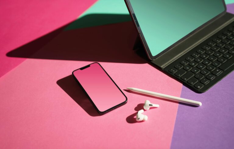 A stylized workspace featuring a smartphone, tablet with keyboard, stylus, and earpods on a vibrant pink and purple backdrop.